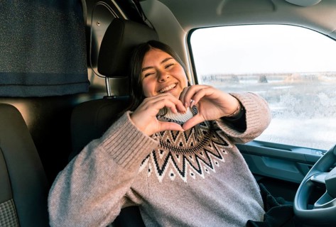 A cheerful Colombian Latina woman in a cozy sweater forms a heart shape with her hands while seated
