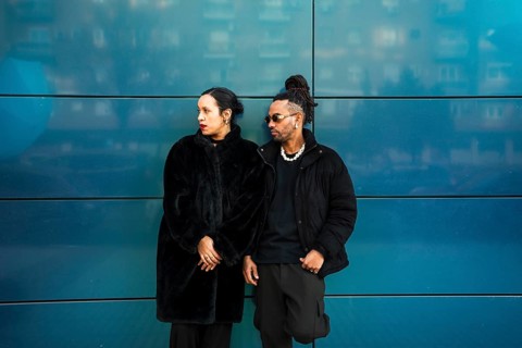 A man and woman from diverse ethnic backgrounds stand against a stylish blue wall. Dressed in