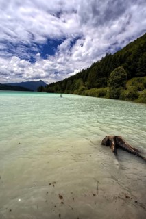 Turquoise-coloured lake with mountain backdrop, cloudy sky and a tree stump in the foreground,