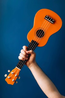 A person holds a bright orange ukulele, standing out against a clean blue background. This vibrant