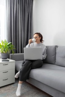 A woman sits comfortably on a sofa, sipping coffee while working on her laptop. She embodies the