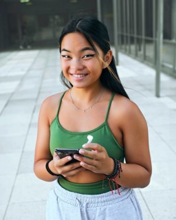 A cheerful teenager with a phone and earbud stands in a bright, outdoor area, wearing a green top