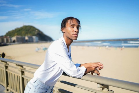 A confident man stands by a beach railing, looking at the camera. The clear blue sky and distant