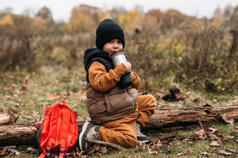 A child sitting on a log, sipping a warm drink in an autumn forest. Wearing a cozy outfit and