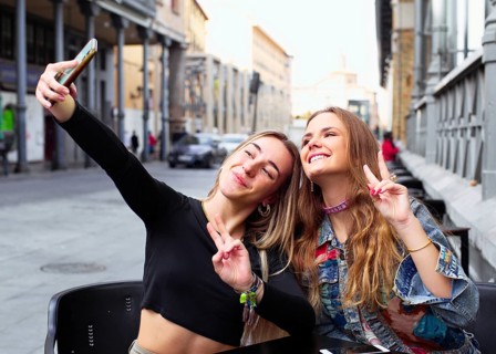 Two teenage girls taking selfies with a smartphone while sitting outdoors. They are smiling,