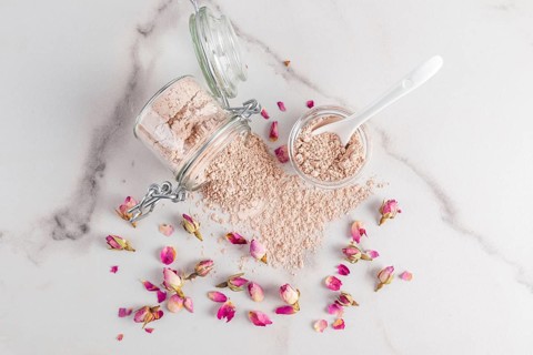 Overhead image showcasing a spilled jar of cosmetic clay powder surrounded by delicate pink rose