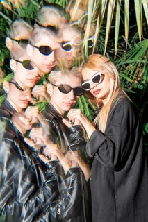 A lesbian couple wearing sunglasses pose in front of lush foliage, showcasing artistic reflections