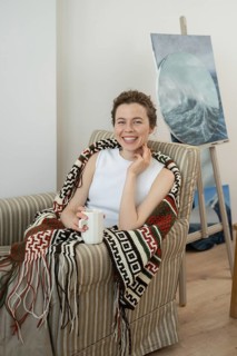 Woman relaxing in a cozy armchair with a colorful blanket, holding a coffee mug. An ocean painting