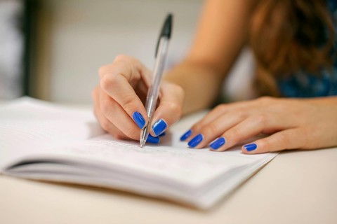 Close-up of a hand with blue-painted nails writing in a notebook. The focus is on the pen and the