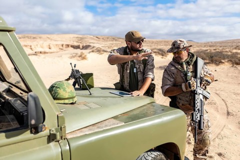 Two military soldiers in tactical gear discuss strategy near an off road vehicle in a desert