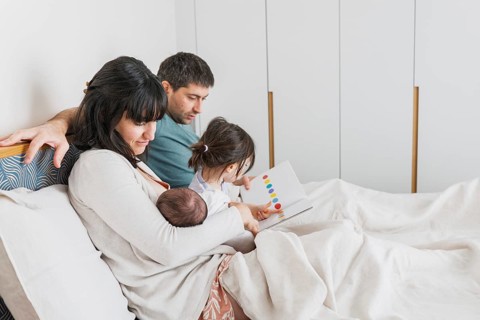 A young family enjoys a cozy moment in their bedroom. The father reads a book with the older child