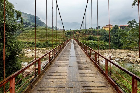 A bridge leads through a tree-lined landscape with rice terraces and ends in the mountains, A