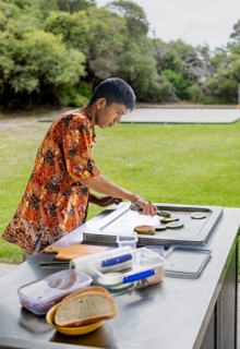 Indonesian man wearing vibrant batik attire prepares food on an outdoor grill. The serene park
