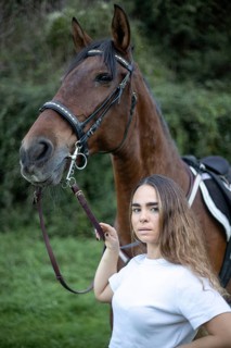 A young woman stands beside a horse in a tranquil outdoor setting. The close-up captures their