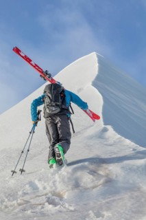 A skier climbs a snowy mountain carrying skis, poles, and backpack, under a clear blue sky. This