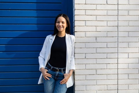 A confident woman with a casual business look, wearing a white blazer and jeans, stands against a