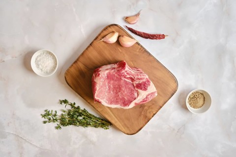 Raw pork loin rack joint on a wooden cutting board. Garlic cloves, dried chili, and fresh thyme are