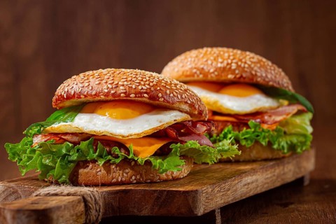 Two juicy burgers sit on a wooden board, each filled with lettuce, bacon, cheese, and fried eggs