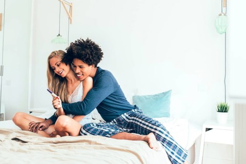 A happy couple sits on their bed, hugging and smiling as they celebrate a positive pregnancy test