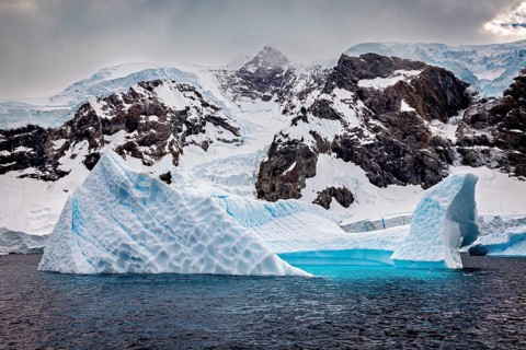 Icebergs with snow-covered mountains in the background under a dramatically darkening sky, icebergs