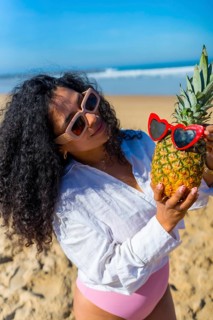 Woman with curly hair and sunglasses standing on a sunny beach, holding a ripe pineapple adorned
