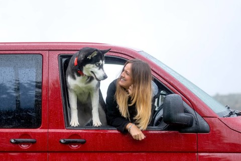 A woman and her dog share a moment through the window of a stationary red van on a rainy day,