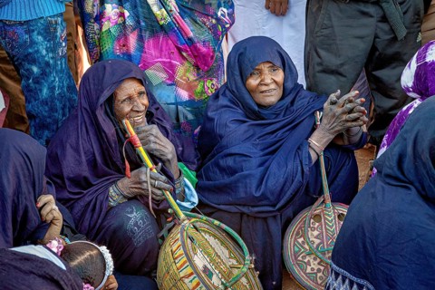 Women in traditional dress sit together holding decorated baskets, surrounded by a group of people,
