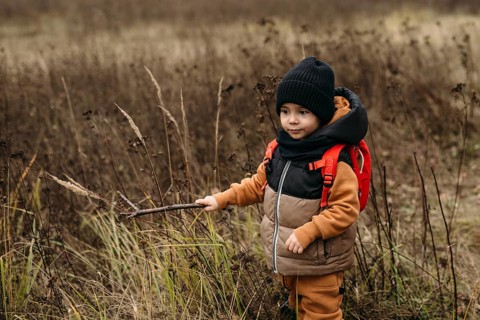 Young boy wearing a warm jacket and backpack explores a field during an autumn walk. He holds a
