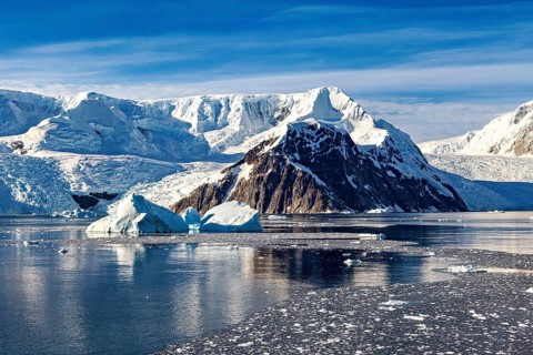 White, clear chunks of ice float in the calm sea in front of imposing white mountains, The