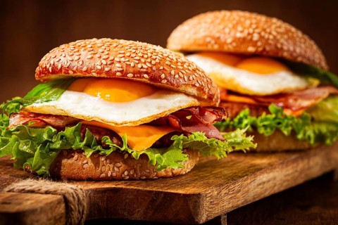 Two juicy burgers sit on a wooden board, each filled with lettuce, bacon, cheese, and fried eggs