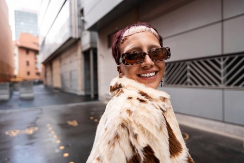 A stylish woman wearing sunglasses and a fur coat smiles confidently in an urban setting. Her chic