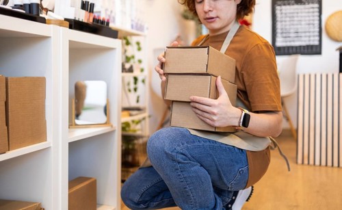 A person kneels in a retail space, organizing cardboard boxes The setting features shelves with