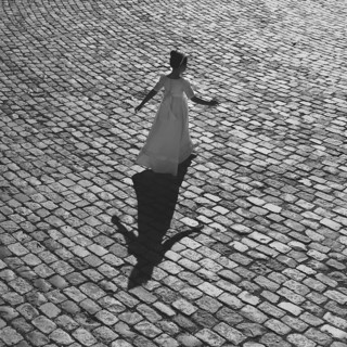 A girl in a First Communion dress walks gracefully on a sunlit cobblestone street, casting a long