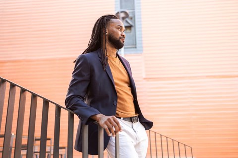 A cuban business man stands by a railing against a modern, orange building backdrop. He wears a