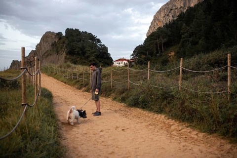 A person walks their dog along a sandy path surrounded by lush greenery and rocky cliffs in Ea,