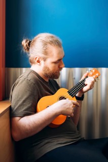 A musician with a beard plays a Mexican ukulele in a cozy setting against a blue wall. The bright