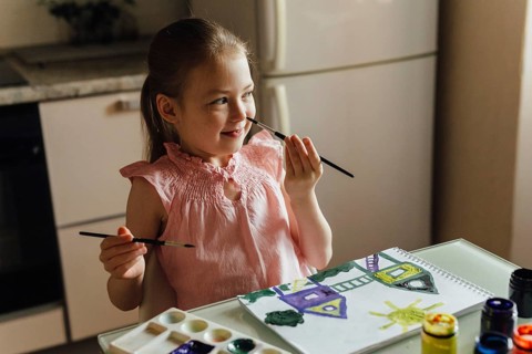 A young girl in a pink dress happily paints a picture with watercolors on paper at a kitchen table.