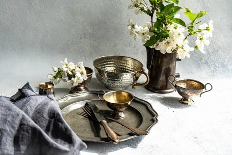 A collection of vintage metal tableware, including cups, a plate, and a jug, is artfully arranged