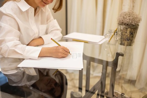 A woman in a white shirt writes on paper at a glass table. Reflections on the surface add depth,
