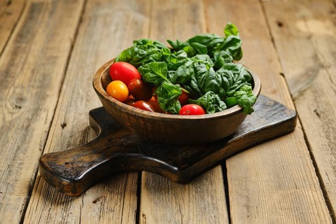 A wooden bowl with red and yellow cherry tomatoes along with fresh green basil leaves. The bowl is