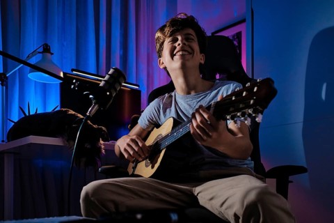 A young musician joyfully strums a guitar in a creatively lit room, surrounded by a microphone and