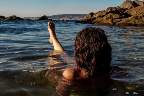 A person with long hair relaxes in the calm, shallow waters by the shoreline. The view includes a