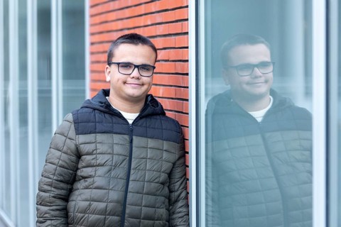 A young man, wearing glasses and a casual jacket, leans against a window at a mental health leisure