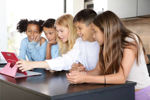 A young woman and four kids gather around a tablet, engaged and smiling in a modern kitchen. The