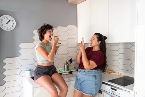 A warm scene of a lesbian couple in a kitchen, one taking a photo while the other enjoys a bite of
