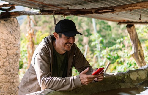 A man in a rural area of Colombia leans on a rustic structure, smiling while using a red smartphone