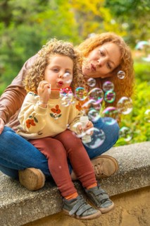 Vertical photo of a curious girl blowing bubbles in the park sitting with her mother