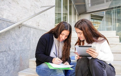 Two students sit on steps outside, discussing and collaborating on assignments One writes on paper,