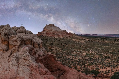 A distant, unrecognizable hiker stands atop a ridged rock formation under a star-filled sky in