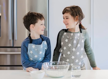 Two children wearing matching star patterned aprons stand at a kitchen counter, smiling at each